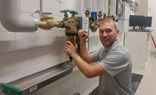Eine kniende Person arbeitet an Rohrarmaturen in einem Technikraum, greift an Ventil und Filter und blickt lächelnd zur Kamera.